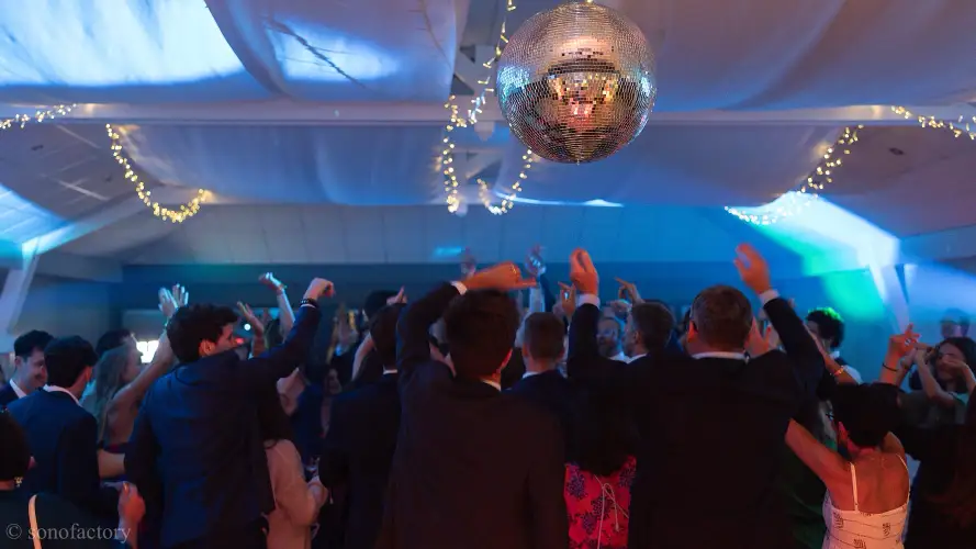 Photographie dynamique d'une piste de danse animée par Sonofactory pour un anniversaire en Bretagne. Une foule enthousiaste danse avec les mains levées sous une grande boule à facettes disco et un éclairage décoratif (déco LED) au plafond. Mixage professionnel et ambiance sur-mesure.